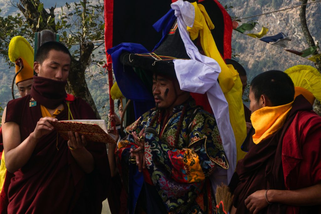 A lama assists the deity lama with scriptures while he holds a brass vajra (a lightning-weapon) and tribu (bell).